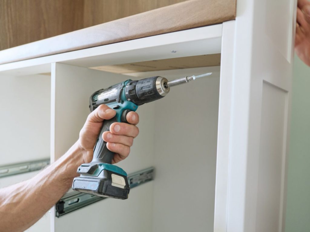 A man operates a drill while installing a cabinet, focused on aligning it properly in a home improvement setting.