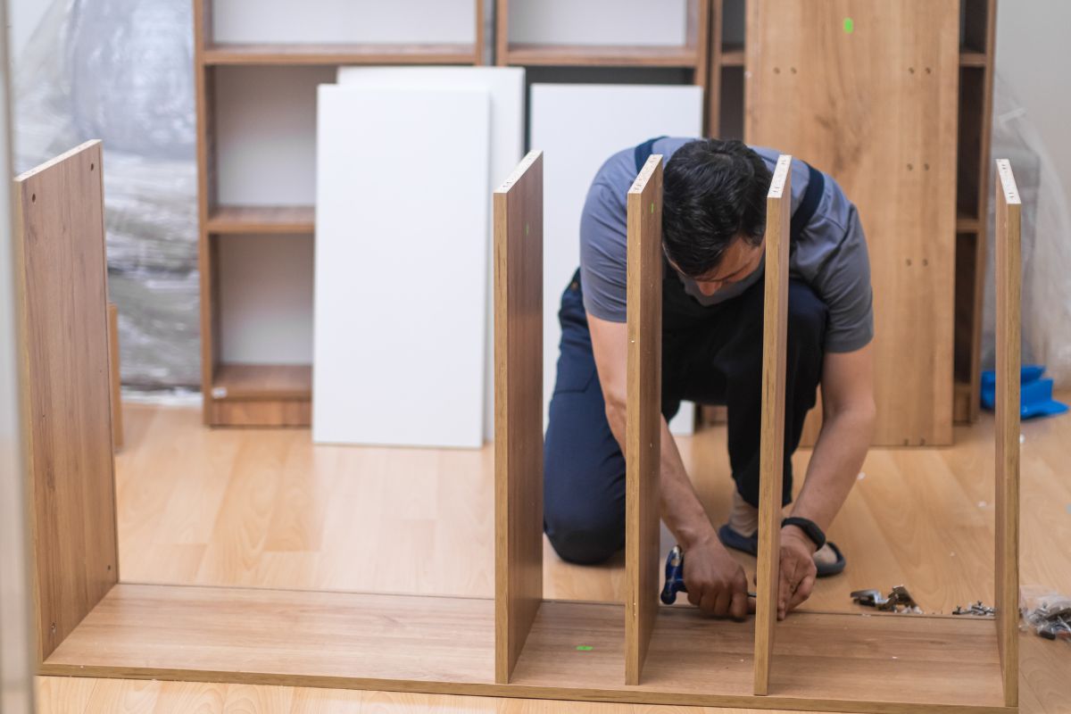 A person assembles a wooden furniture piece on a wooden floor, with various wooden components and shelves in the background.