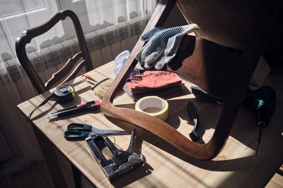A wooden chair frame on a table surrounded by various tools, including scissors, a tape measure, gloves, and a stapler.