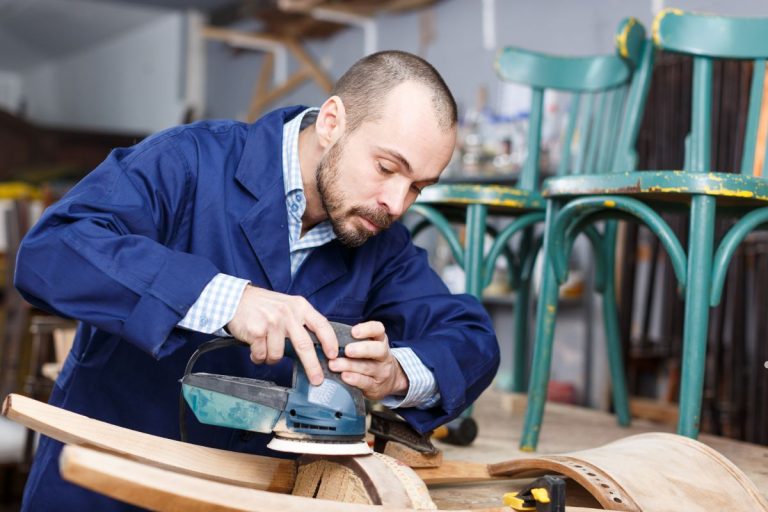 A craftsman in a blue work jacket uses an electric sander on a wooden piece in a workshop surrounded by colorful chairs.