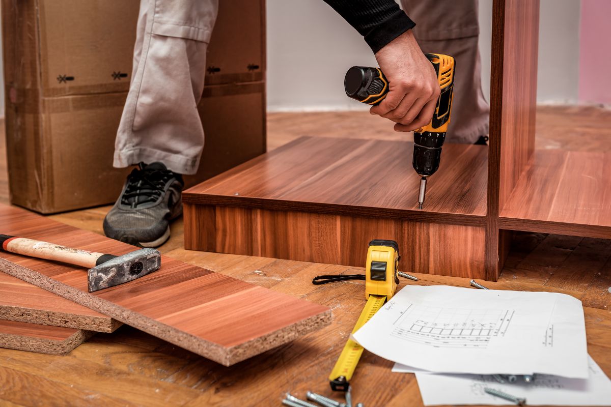 A person using a power drill to assemble furniture, surrounded by tools, including a hammer, measuring tape, and assembly instructions.