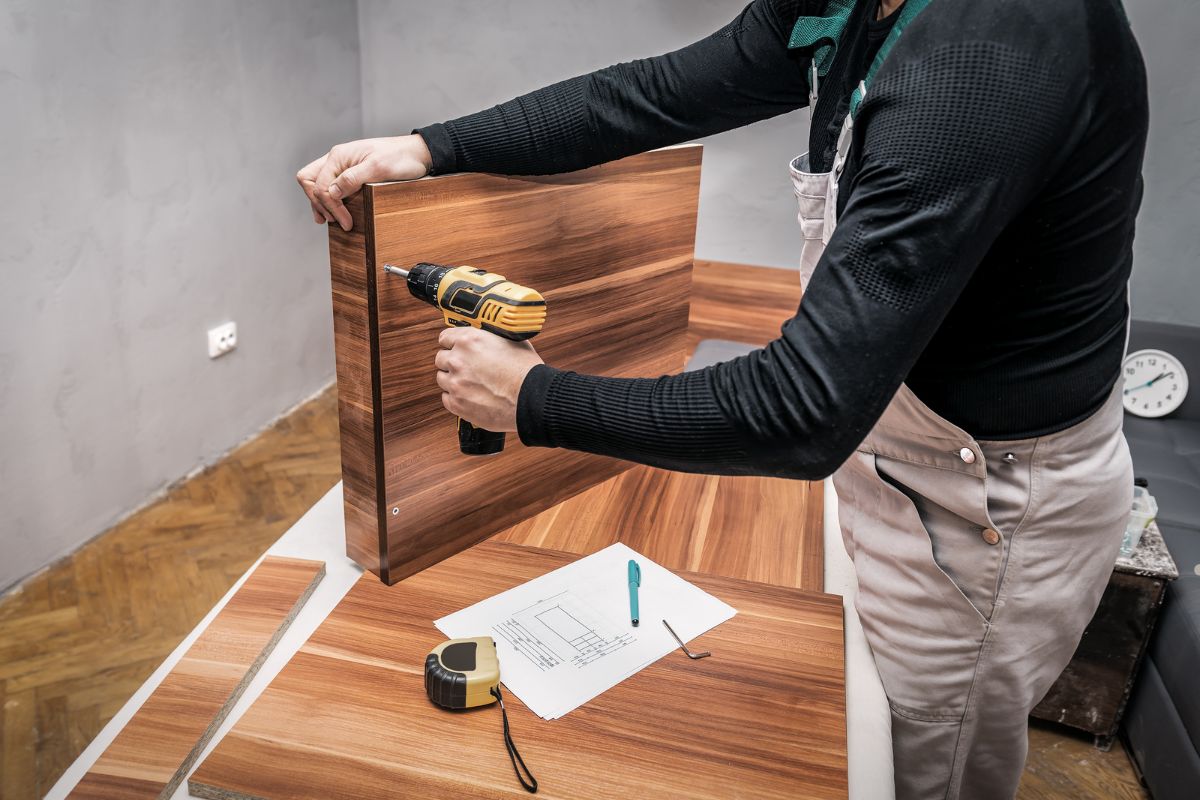 A person using a power drill to assemble wooden furniture, surrounded by pieces of wood and a blueprint on a table.