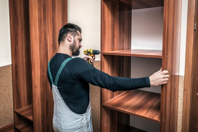 A person in a gray apron is assembling wooden shelves using a yellow cordless drill in a room with wooden paneling.